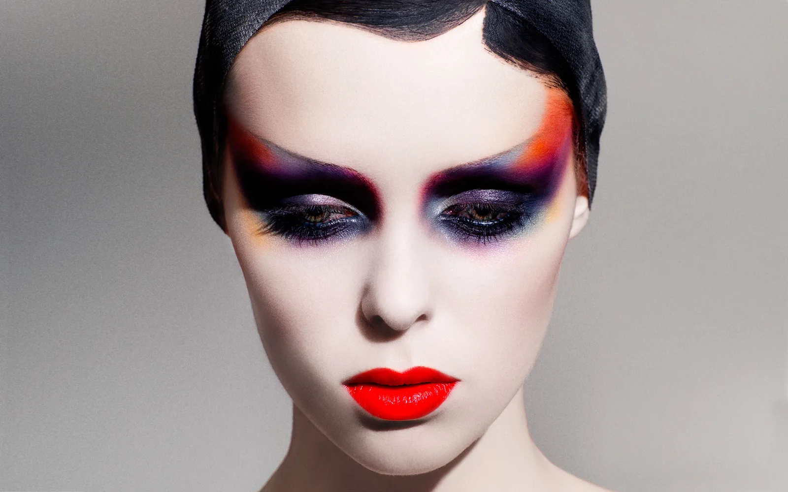 Portrait beauté retouché : visage pâle, fards violets, bleus et orangés, lèvres rouges, sur fond gris.