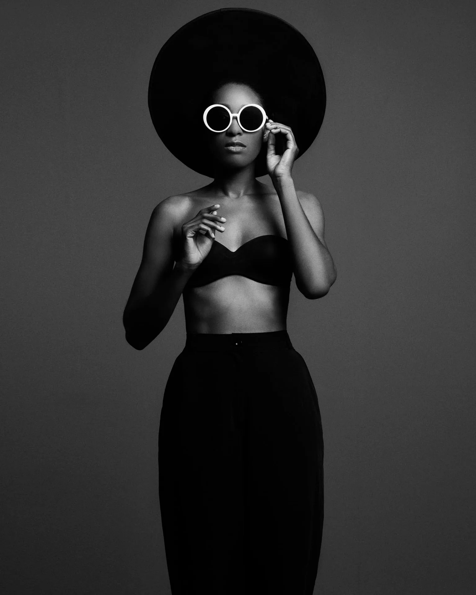 Photographie noir et blanc : femme au grand chapeau et aux lunettes rondes blanches, sur fond gris.
